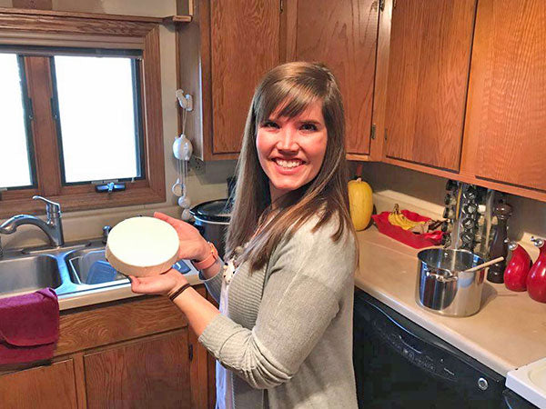 A woman stands in a kitchen and holds up a wheel of homemade cheese for the camera