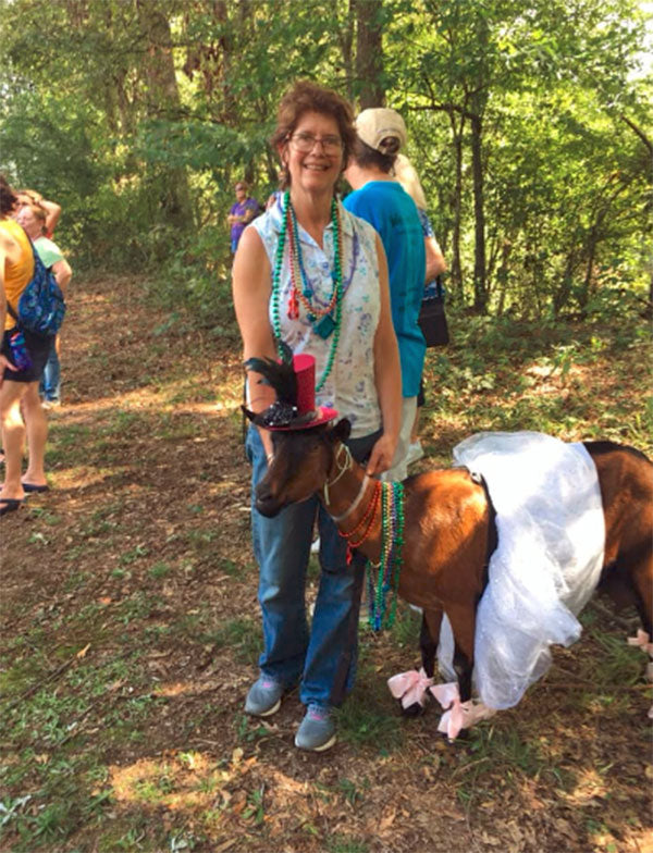 A woman stands with a goat that is dressed in a tutu, beads, and a pink hat with black feathers