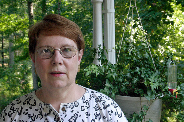 A middle aged white woman with short reddish brown hair, metal rim galsses, and a white shirt with leopard print stands in front of a hanging plant with ivy.