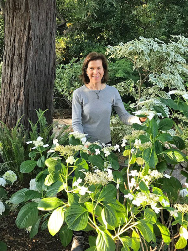 A woman in a light gray shirt smiles at the camera while standing in a garden with white flowers