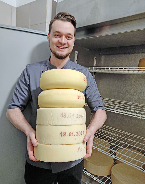 A younger white male holds five large wheels of cheese while smiling at the camera. He is wearing a gray shirt and black pants and is standing in front of what appears to be an open industrial refrigerator.