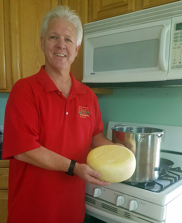 A white male with short white hair wearing a red polo shirt holds a wheel of cheese while standing in front of his stove. There is a stainless steel pot on the stove.