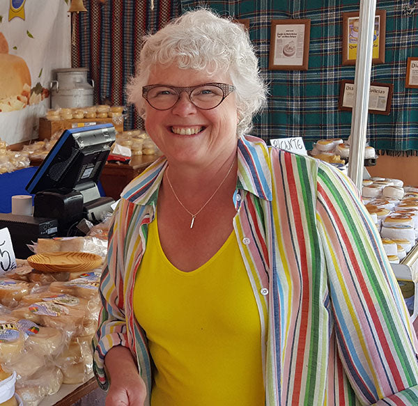 A woman with short white hear and glasses wearing a yellow top with an open striped button up over it smiles at the camera in front of cheese samples